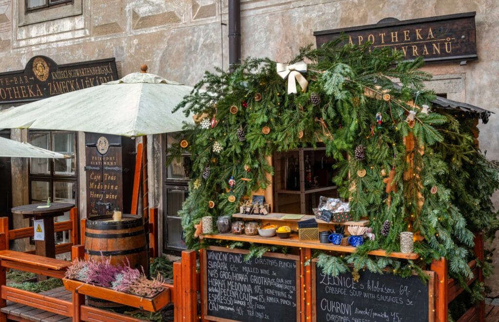 A charming Christmas market stall in Český Krumlov, decorated with fresh pine branches, dried oranges, pinecones, and festive ornaments. The wooden booth offers traditional holiday treats, including mulled wine, honey wine, and Christmas sweets. The cozy setting is enhanced by a rustic outdoor seating area, inviting visitors to enjoy the festive atmosphere.