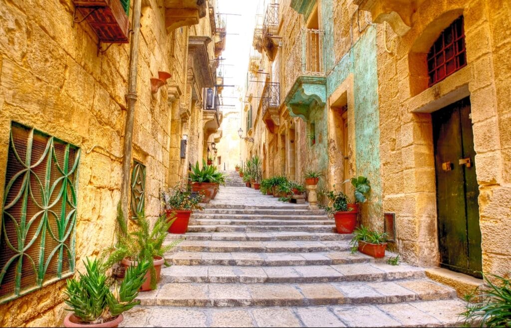 Scenic alleyway features stone staircase flanked by historic limestone walls, potted plants, and traditional colored doors in Gozo